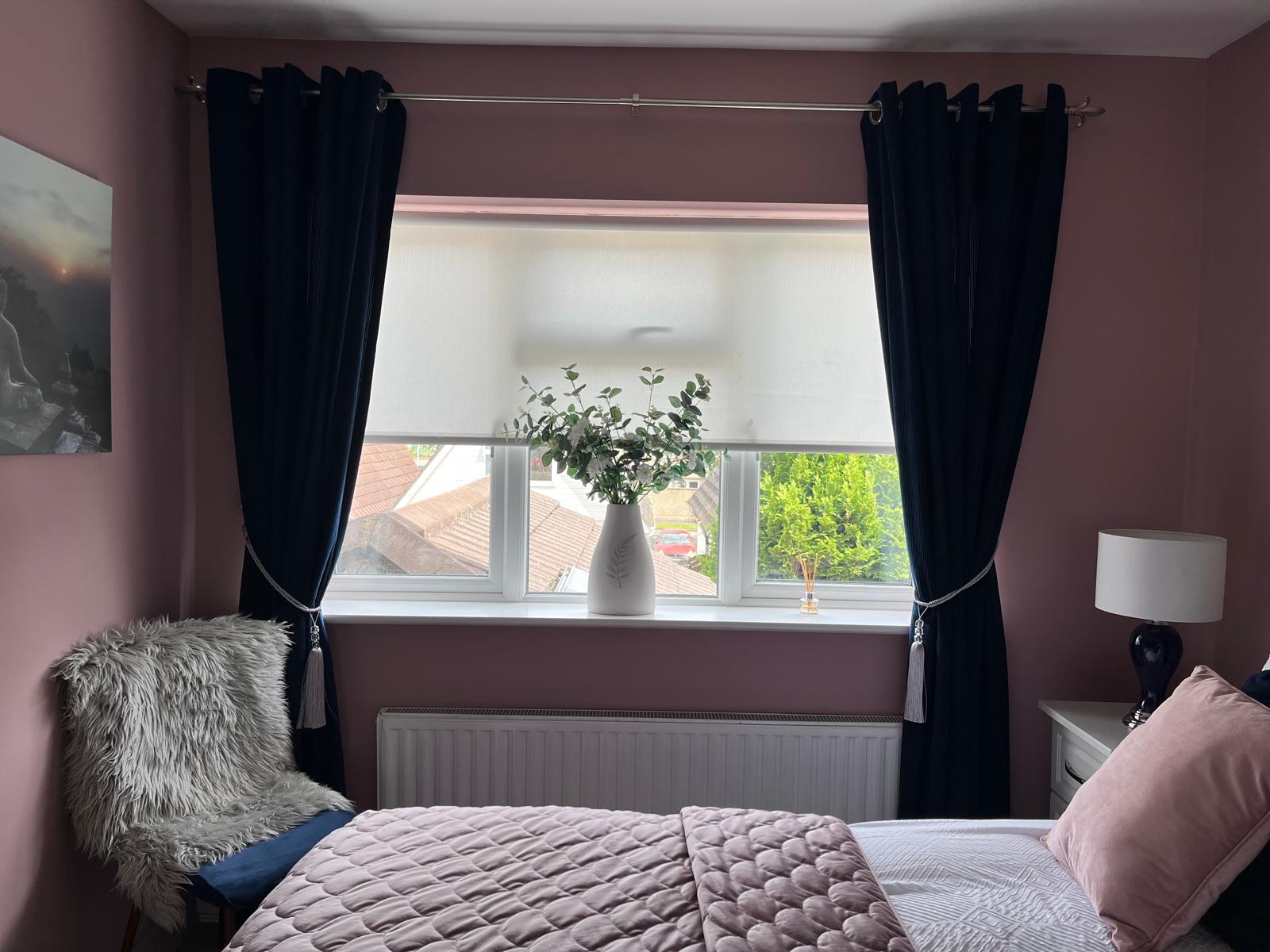 a pink bedroom, window surrounded by navy, ribbed curtains and silver tie backs. There is eucalyptus in a vase on the window sill. A chair sits in corner with faux sheep skin on. There is calming artwork on the walls. a Pink throw on the bed.